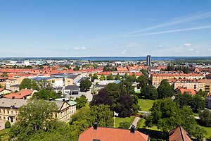 Flygbild över en stad med röda tak, parker och många träd mellan byggnaderna. I bakgrunden syns vatten och horisont under en klar blå himmel.