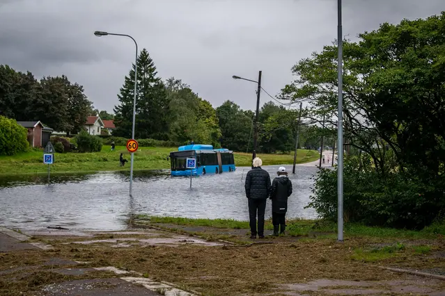 Två personer står vid en översvämmad väg där vatten täcker gatan. En blå buss har fastnat i vattnet och trafikskyltar sticker upp ur översvämningen.