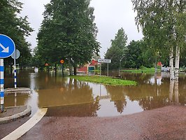 Översvämmad väg i Gävle efter kraftigt skyfall. Vatten täcker gatan vid infarten mot Hemlingby medan träd, trafikskyltar och byggnader syns i bakgrunden.