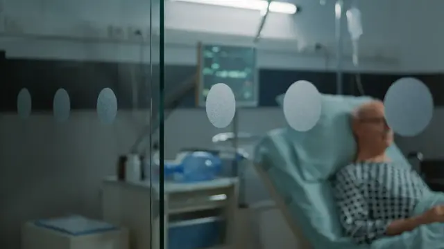 En person ligger i en sjukhussäng, bakom en glasvägg.